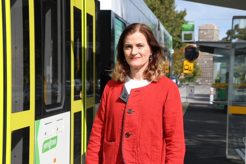 A woman in a red jacket stands on a tram stop next to a Melbourne tram.