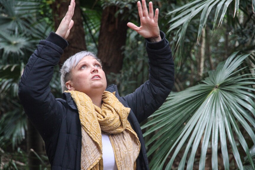 A woman with short white hair and a big scarf around her neck raises her arms to the sky while surrounded by green palm fronds