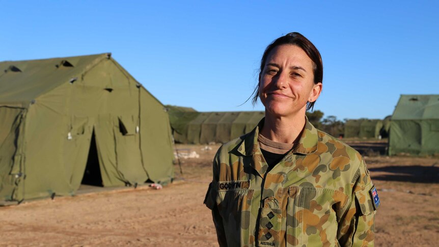 A woman in army greens stands proudly and smiling against a backdrop of army green tents