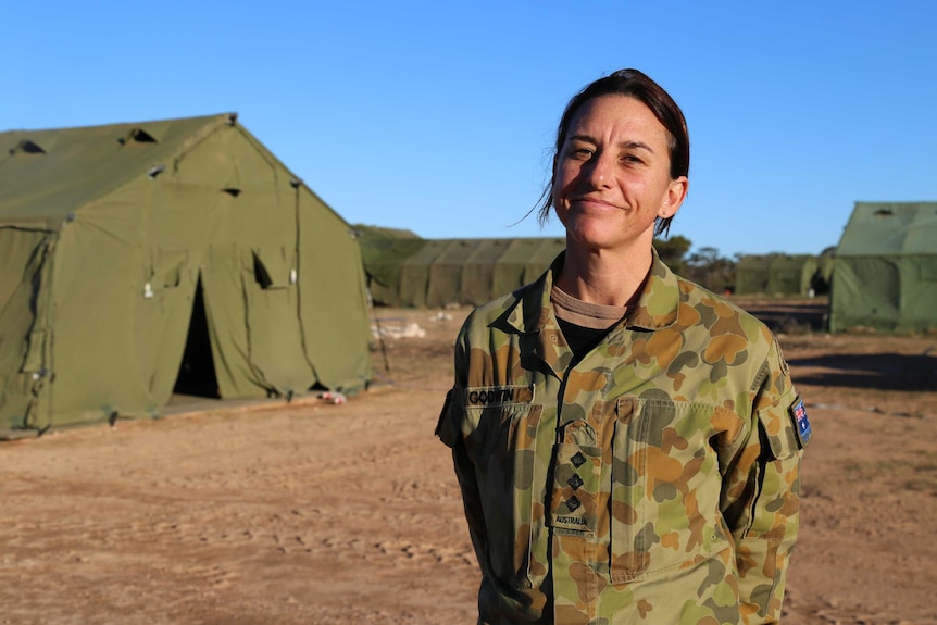 A woman in army greens stands proudly and smiling against a backdrop of army green tents