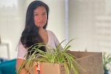 A woman with long dark hair wearing a white t-shirt holds a cardboard box with a leafy plant inside.
