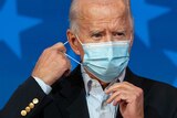 Joe Biden stands in front of microphones and adjusts the elastic loops of his face mask around his ear