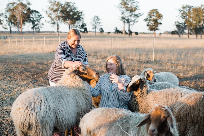 Two women in a paddock petting a group of sheep.