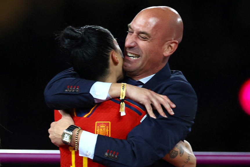 Ein lächelnder Mann im Anzug umarmt eine Frau in Fußballuniform