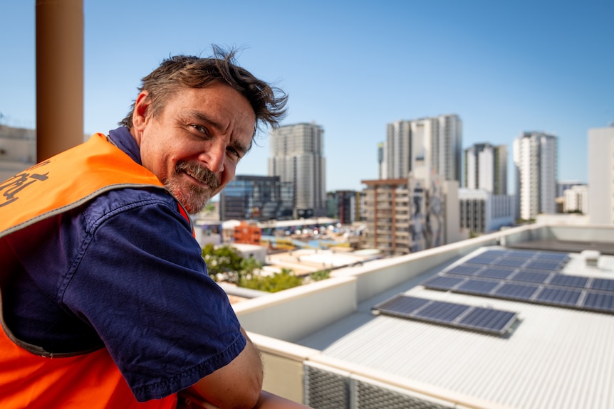 A man in a hi-vis vest looking at the camera while leaning on a balcony, with tall buildings in the background.