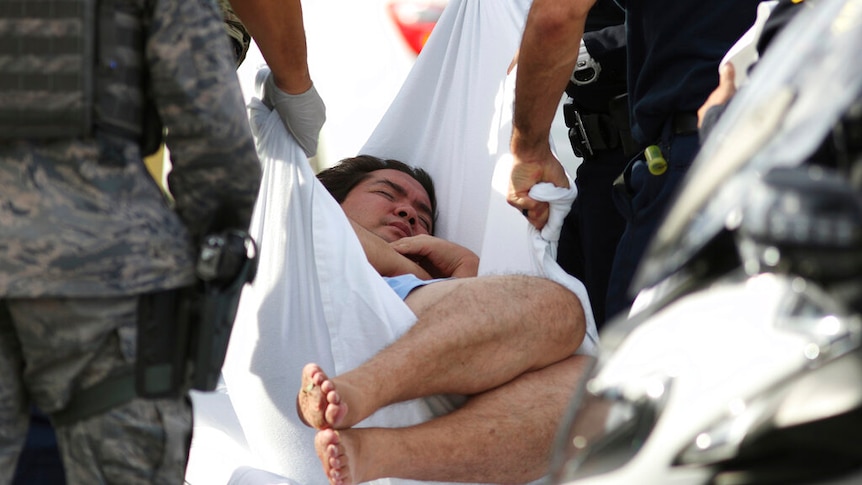 A man holds his arms to his chest as he is wrapped in a white sheet and is placed on an ambulance stretcher.