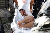 A man holds his arms to his chest as he is wrapped in a white sheet and is placed on an ambulance stretcher.