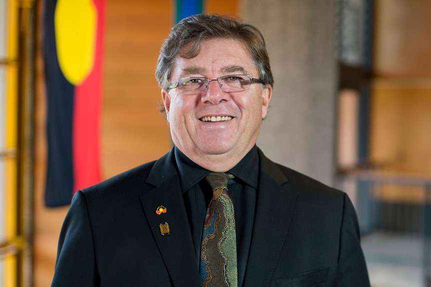 Professor John Maynard in a black suit stands in front of an Aboriginal flag.