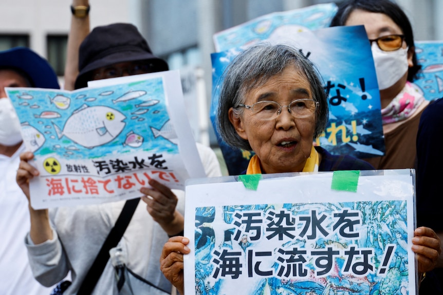 An elderly woman stands holding up a laminated sign that says "Don't throw radiation contaminated water into the sea"