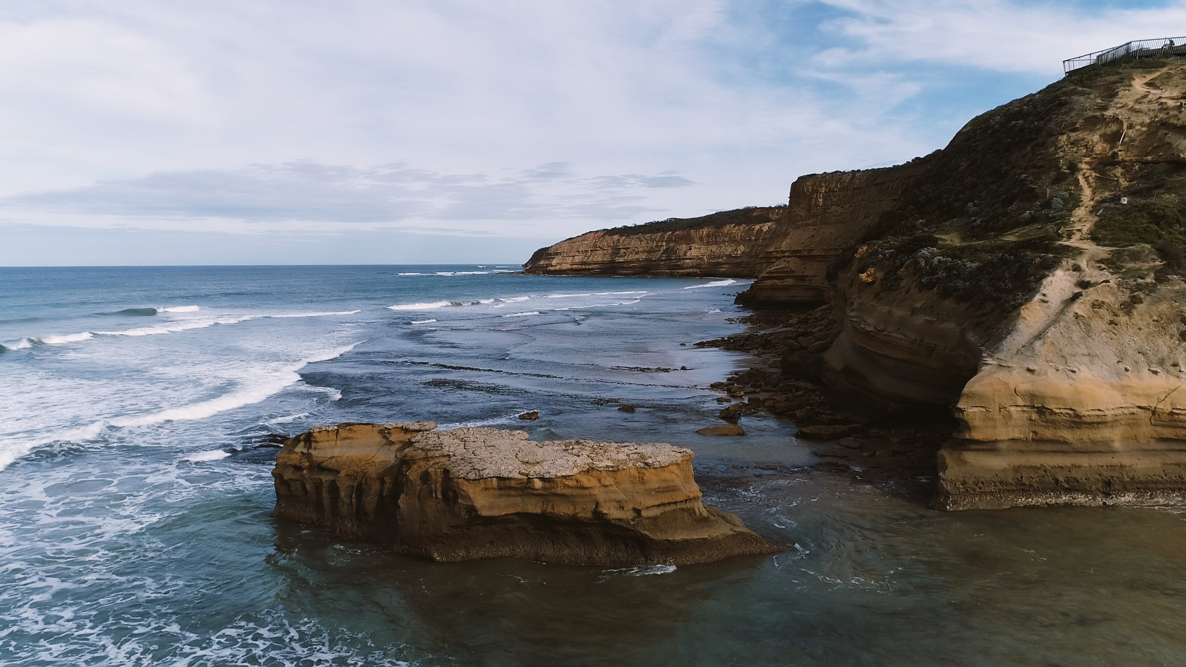 Waves approach a large rock that juts out of the water beside a rocky sea cliff on a partly cloudy day.