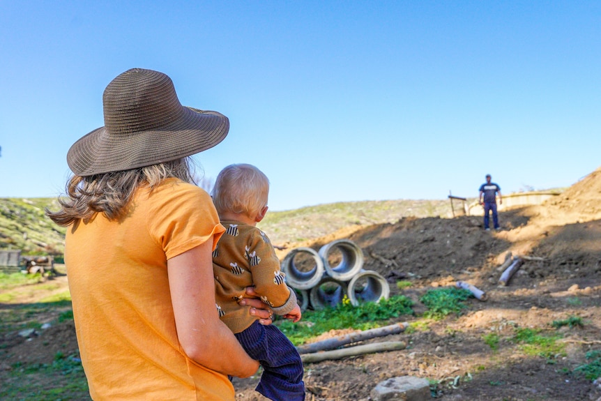A woman wearing a broad brim hats hols a baby girl on her hip as she overlooks a construction site on a sunny afternoon.