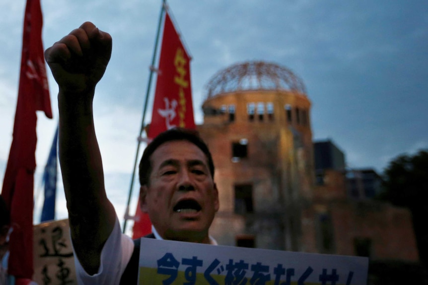 Close up view of a Japanese protester, fist raised and holding a sign, standing in front of the Atomic Bomb Dome in Hiroshima.