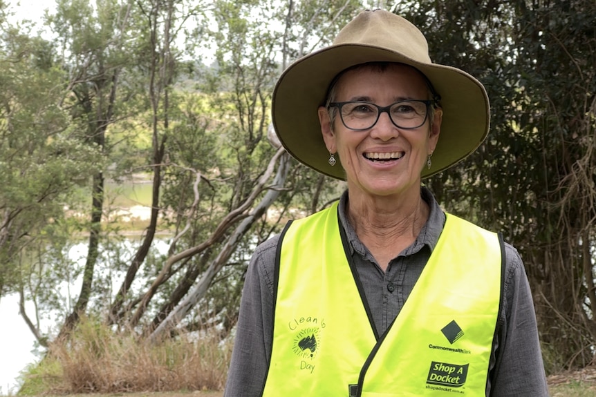 A woman in a hat and glasses and wearing a high-vis vest smiles at the camera, a waterway and greenery behind
