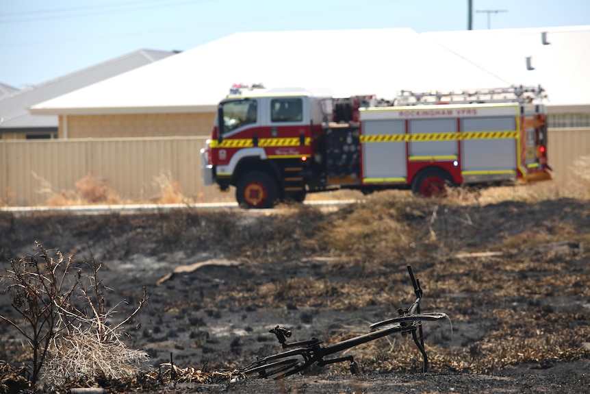 A burnt bicycle lies amid the scorched bushland with a firetruck in the background