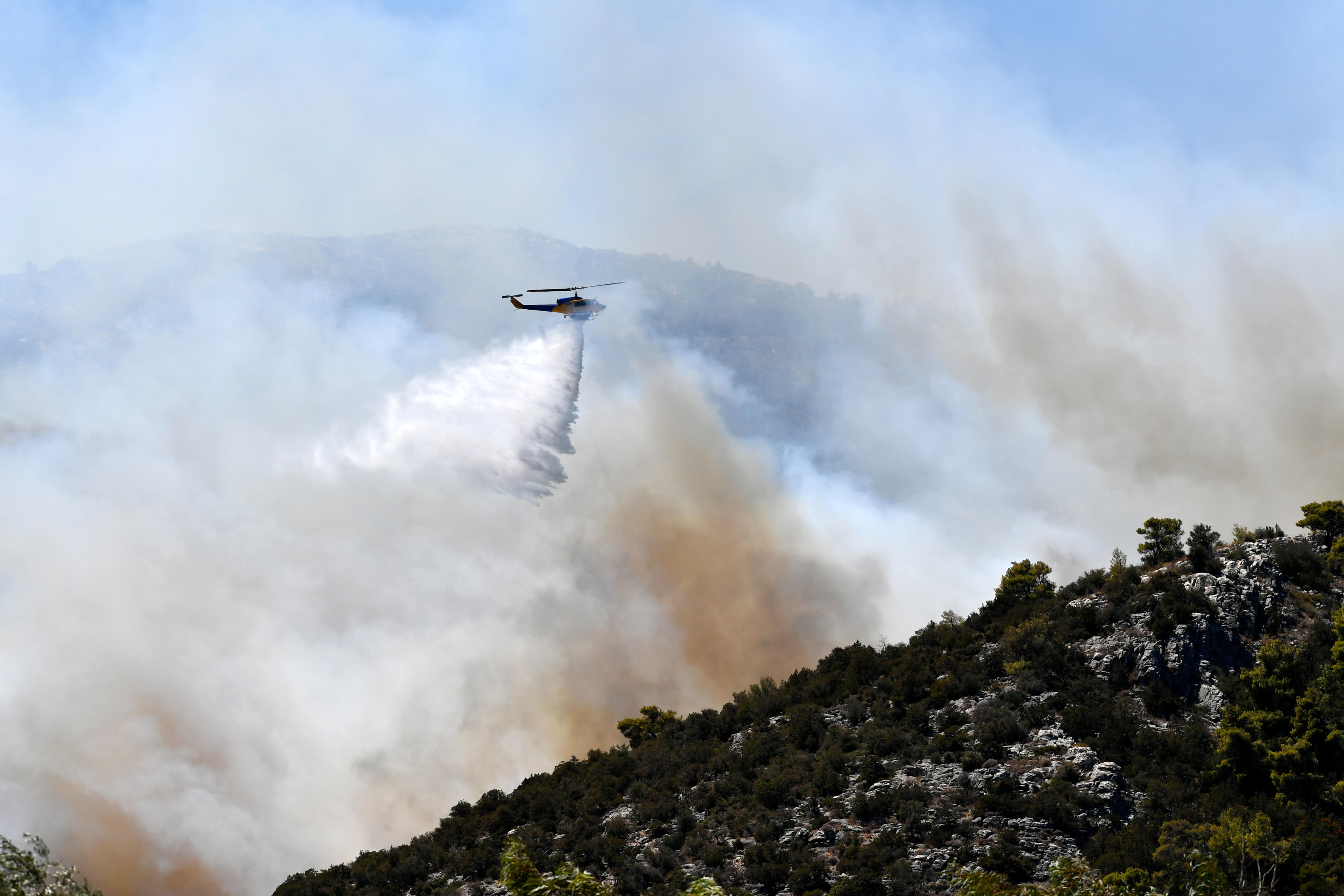 A helicopter drops water over a shrubby hill, while smoke rises in the background.