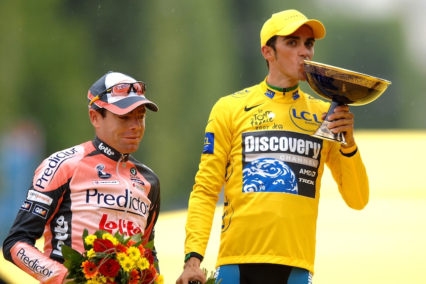 Cadel Evans holds flowers standing next to Alberto Contador who kisses a trophy while wearing yellow