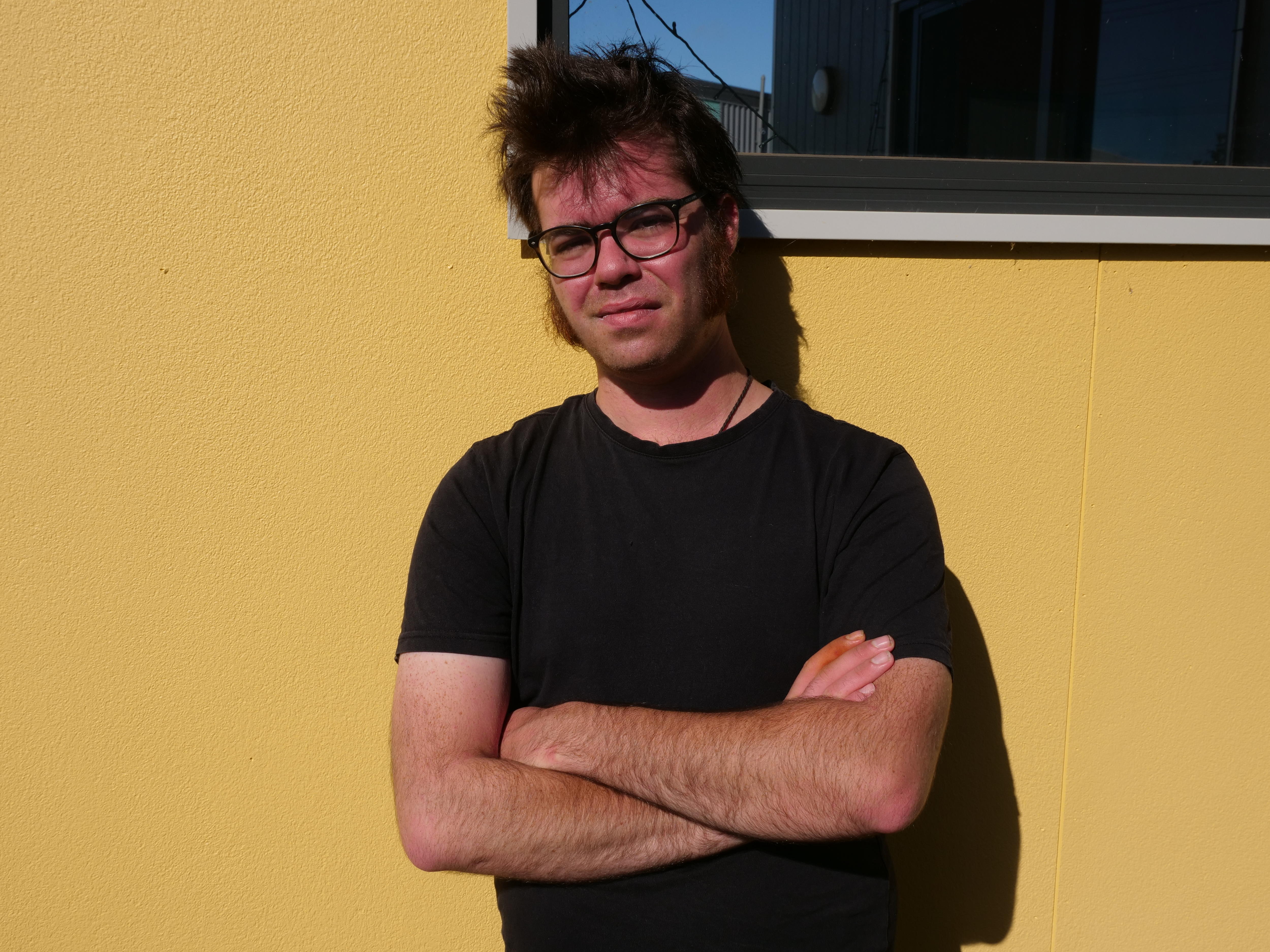 a man in a black t shirt leaning on a yellow wall, his arms are crossed 