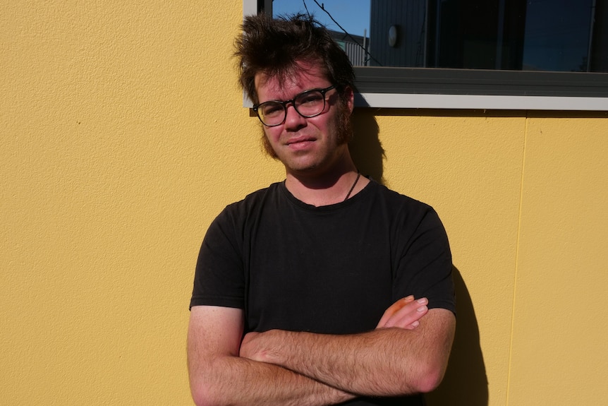 a man in a black t shirt leaning on a yellow wall, his arms are crossed 