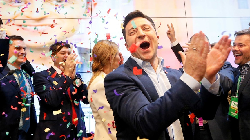 Volodymyr Zelenskiy laughs and claps his hands on the stage as confetti falls from above.