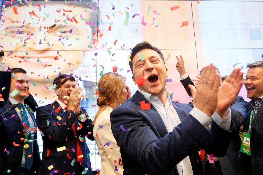 Volodymyr Zelenskiy laughs and claps his hands on the stage as confetti falls from above.