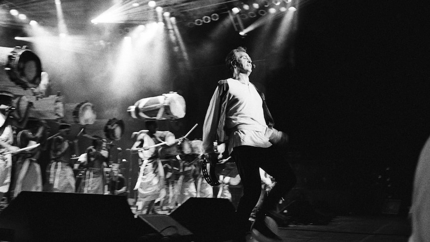 A black and white photo of Peter Gabriel on stage dancing. Behind him are many drummers.