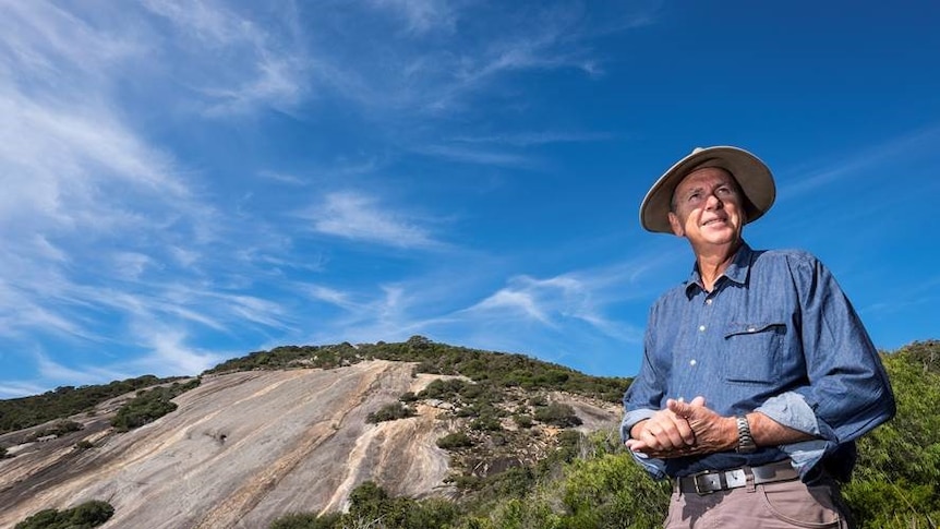 A man in a wide-brimmed hat standing in front of a granite outcrop.