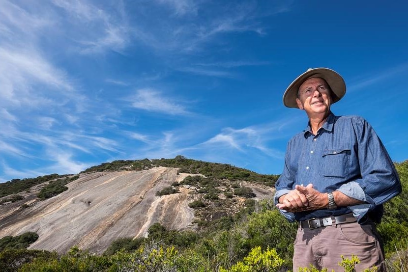 A man in a wide-brimmed hat standing in front of a granite outcrop.