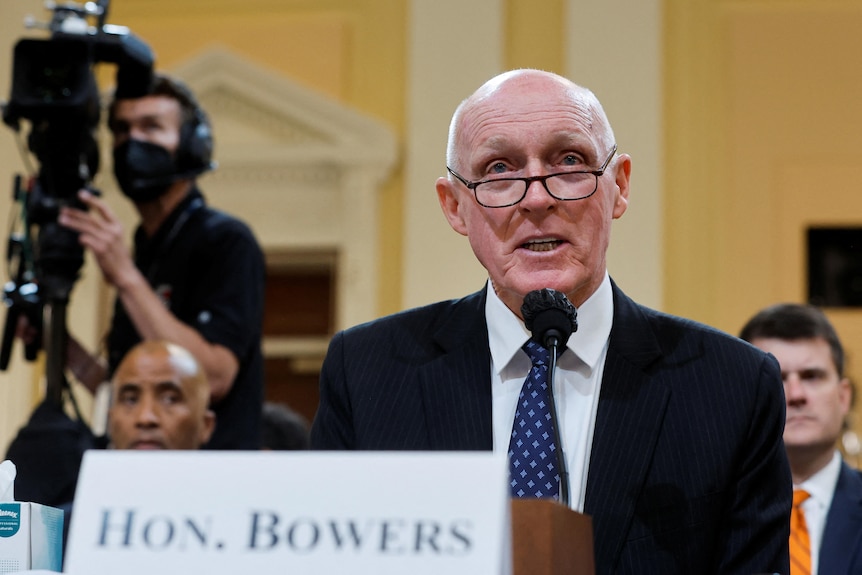 Rusty Bowers in a suit and blue tie testifying at a microphone, with camera in background