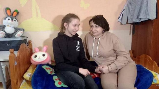 A girl in her early teens sits on a bed smiling and holding hands with a middle-aged woman.