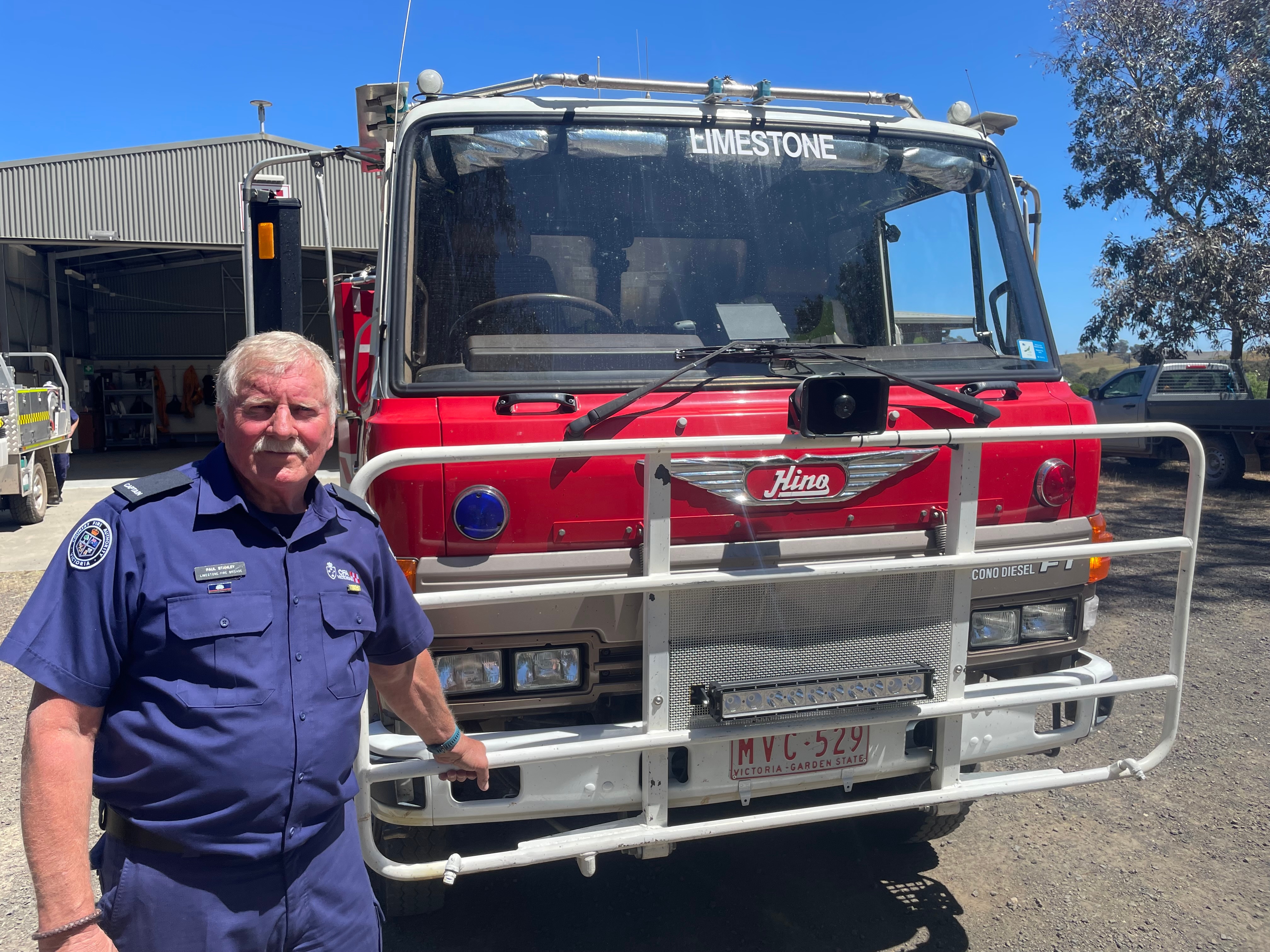 A man wearing a navy blue CFA uniform stands in front of a red firetruck.