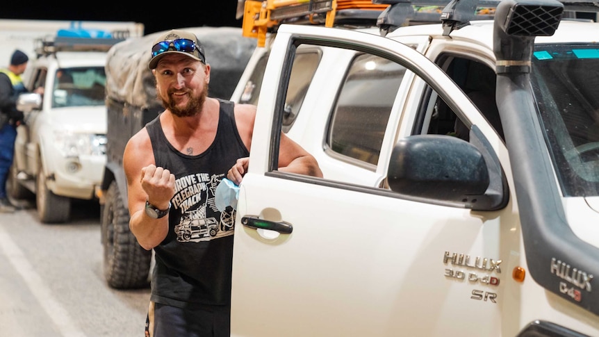 a man in a black singlet raises his fist happily as he stands outside his ute in a line up of cars