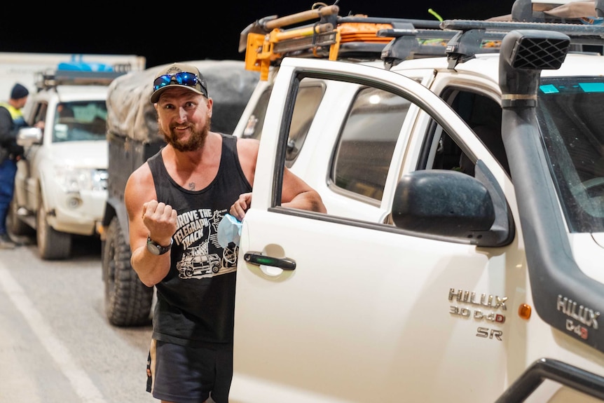 a man in a black singlet raises his fist happily as he stands outside his ute in a line up of cars