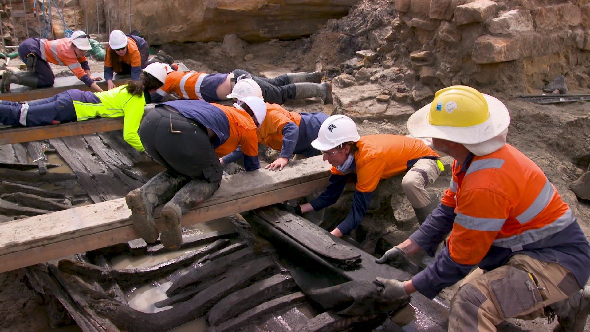 Archaeologists wearing high-vis and protective gear working together to excavate colonial-era boat.