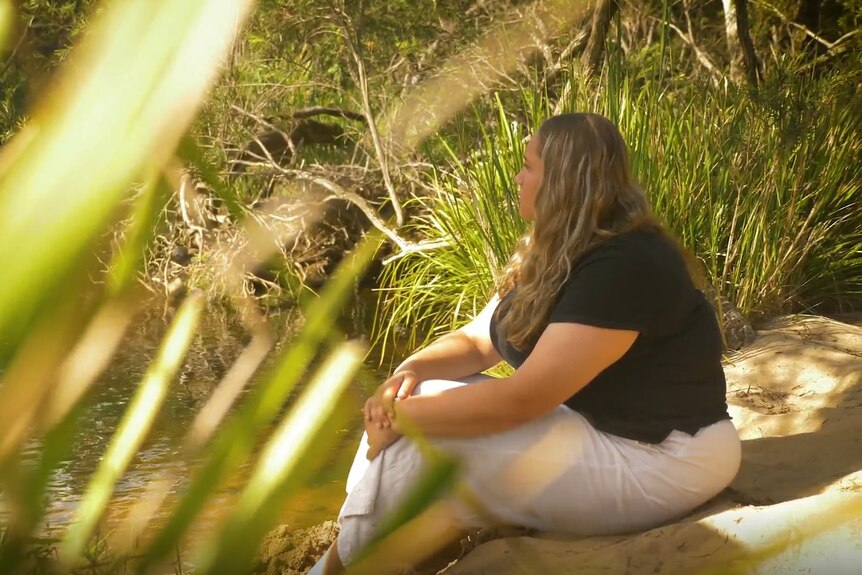 照片中，一名身穿黑色衬衫和白色裤子的金发女孩坐在河岸上，双脚浸在水中。 