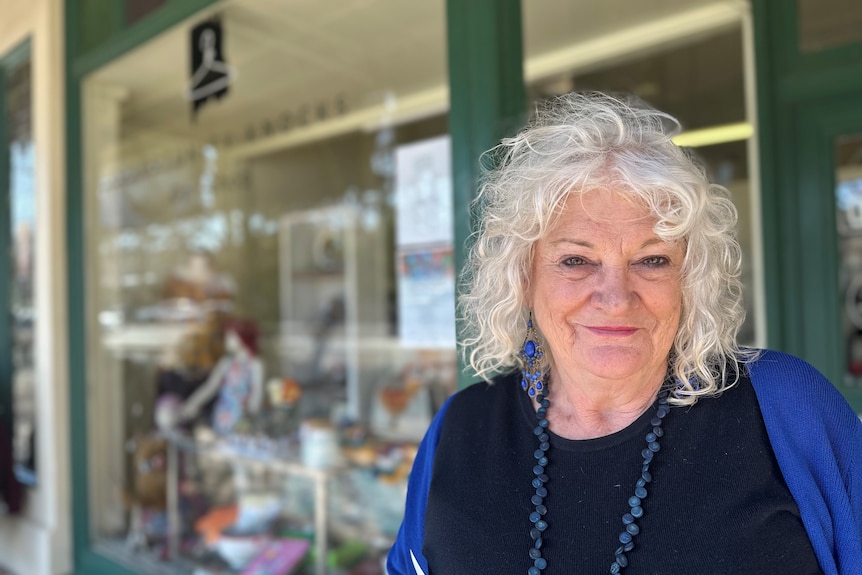 A grey-haired woman smiles in front of a sjop front window. 