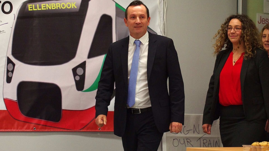 Mark McGowan walks towards the camera wearing a suit with a woman behind him and a picture of a train on the wall.