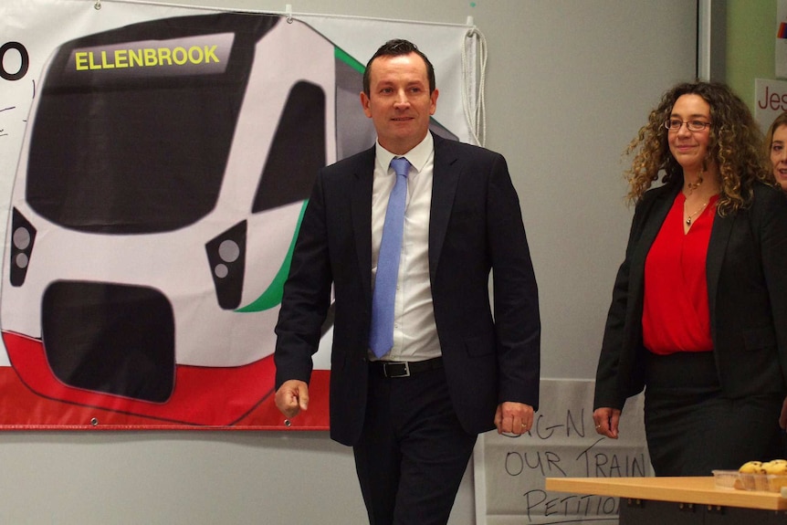 Mark McGowan walks towards the camera wearing a suit with a woman behind him and a picture of a train on the wall.
