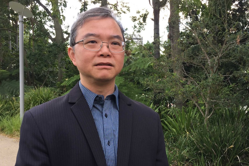 A man with large rectangular glasses and thin-rimmed frames and greying hair speaks in a park.