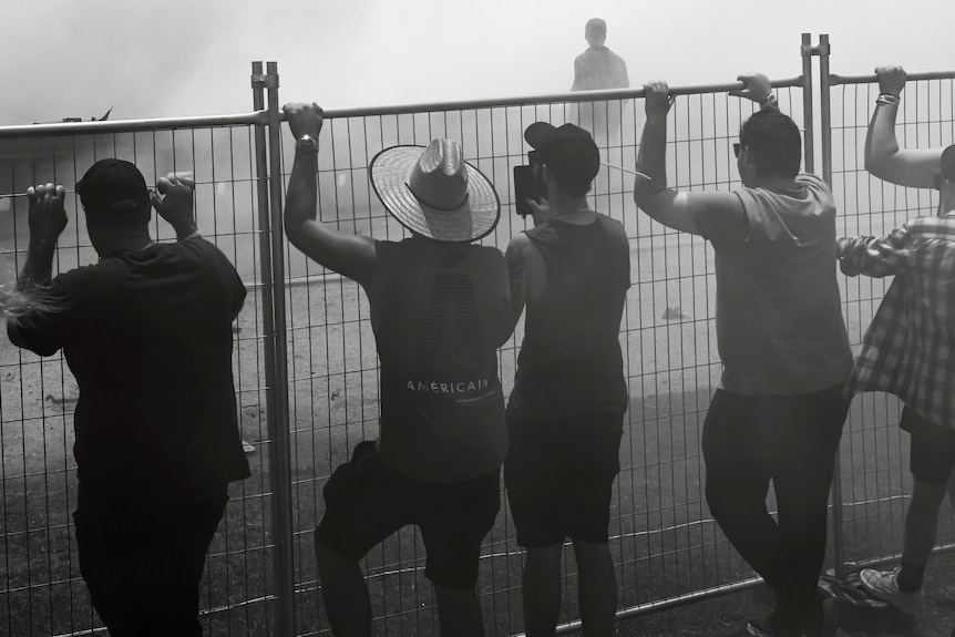 A black and white photo of a crowd staring in the smoke of a burnout from behind a fence.