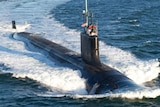 A submarine moves through the water causing a massive wake behind it, some crew visible above deck.