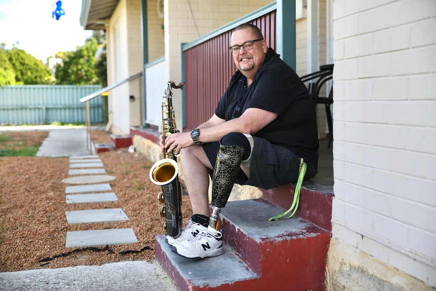 Andrew Fairbairn sitting on step playing sax, his left leg has a black, patterned prosthetic and he is wearing shorts.