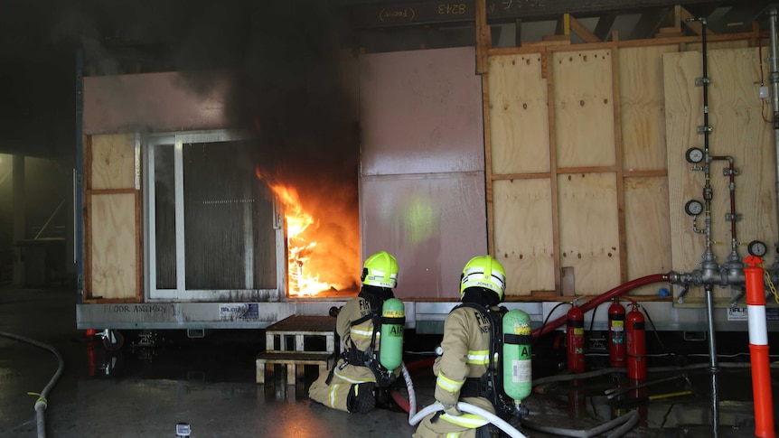 Two fire fighters crouch outside a plywood structure in another building as flames and black smoke fill the wooden structure