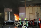 Two fire fighters crouch outside a plywood structure in another building as flames and black smoke fill the wooden structure