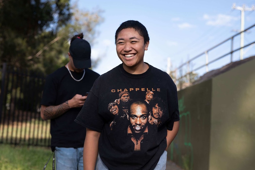 Jada wearing t-shit with David Chappelle's name and face, man looking down at mobile phone in background.