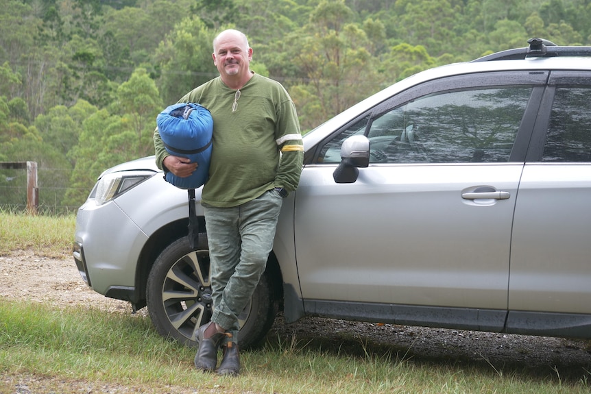 A man leaning on a car with a sleeping bag under his arm.