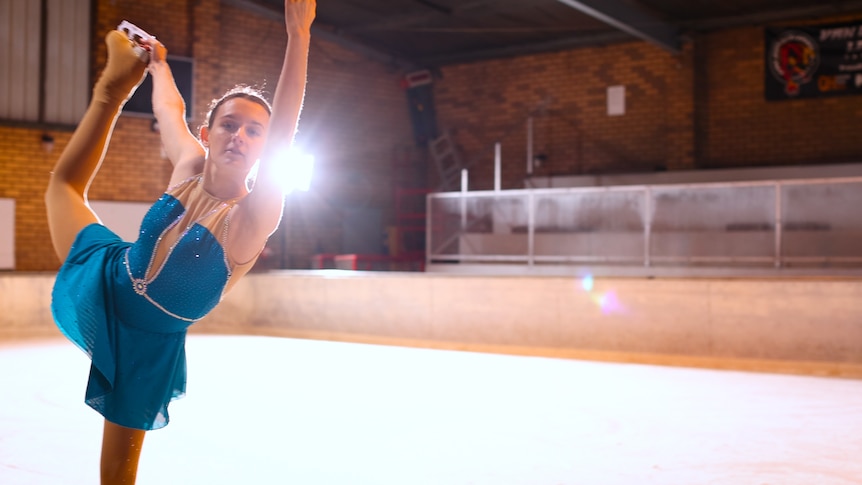 A woman in a blue sparkly leotard is ice skating, holding one leg behind her head