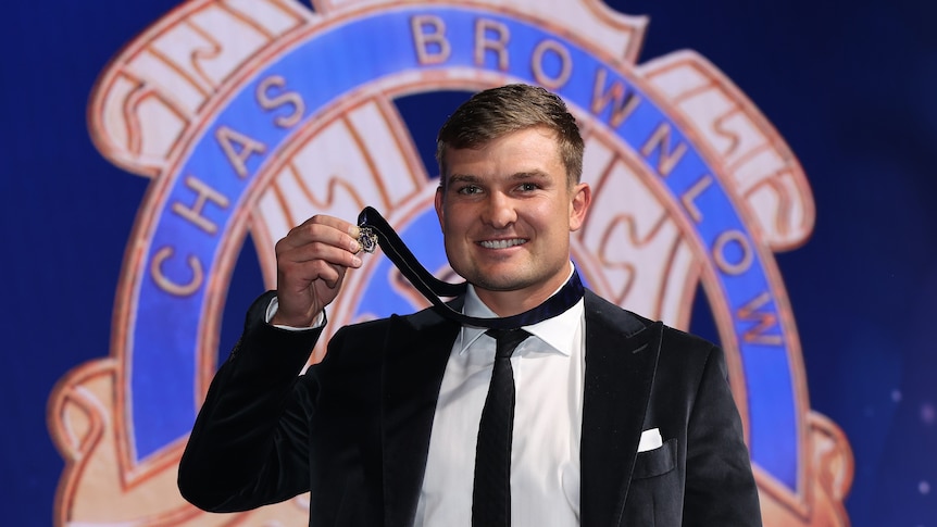 A Port Adelaide AFL player smiles at the camera and holds the Brownlow medal as it sits around his neck.