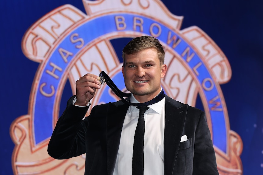 A Port Adelaide AFL player smiles at the camera and holds the Brownlow medal as it sits around his neck.