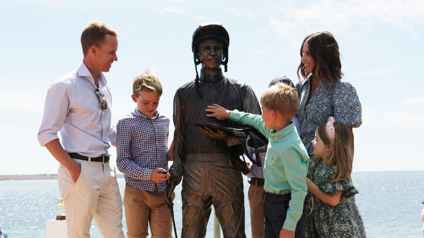 Man and son on left and woman on right with two boys and girl looking at bronze life size jockey statue in the middle