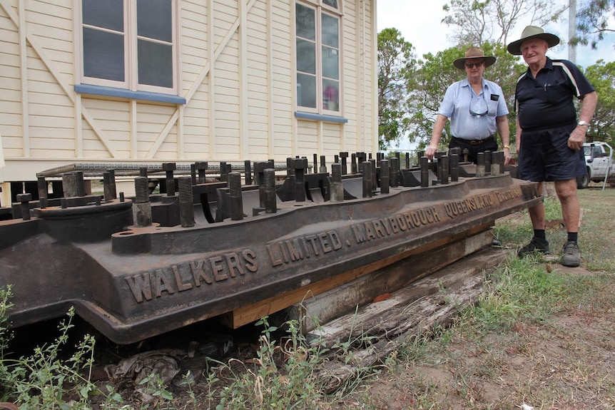 Two men stand behind the block of  a massive vintage engine removed from a sugar mill